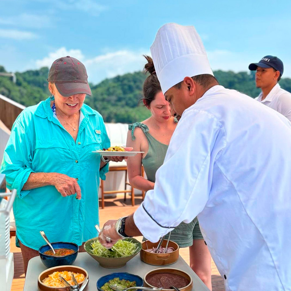 Chef in a white uniform and tall hat serves salad at an outdoor buffet as guests wait with plates under a blue sky.