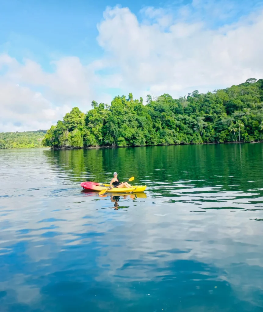Person kayaking in a yellow boat - Kontiki Small Ship Expedition Person kayaking in a yellow boat on calm water near a lush green shoreline with blue sky and white clouds behind.